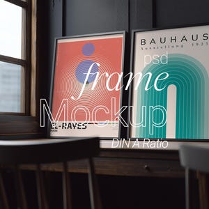 May include: Two framed art prints with black frames are displayed on a wooden surface near a window. One print features a red and orange design with blue and purple accents. The other print has a teal and white design with the text "BAUHAUS Ausstellung 1923".