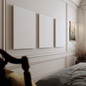 Può includere: Tre tele bianche vuote di diverse dimensioni sono montate su una parete beige chiaro. Il muro presenta modanature decorative e un piccolo dipinto incorniciato. Una sedia in legno scuro e un letto con un copriletto grigio sono in primo piano.