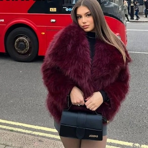 May include: A deep burgundy faux fur coat worn with a black top and a black handbag. The handbag is rectangular with a top handle and a gold logo. A red double-decker bus is visible in the background.