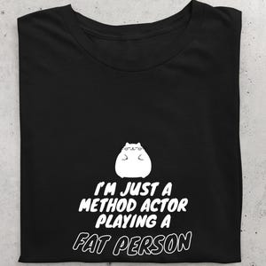 Può includere: T-shirt nero con un gatto bianco dei cartoni animati con gli occhiali. Il testo recita "I'M JUST A METHOD ACTOR PLAYING A FAT PERSON" in un carattere giocoso. La maglietta è piegata, mostrando il design.