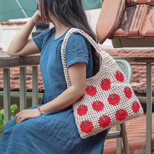 May include: A beige crochet tote bag with a pattern of red tomatoes and green stems. The bag is held by a person wearing a blue dress. The text "@tinycrochet" is visible on the bag.