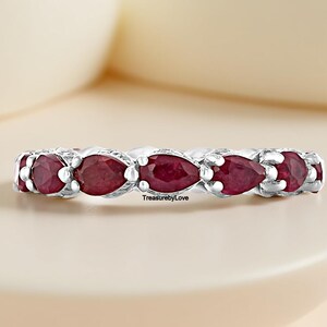 Può includere: Un anello d'argento con una serie di pietre preziose rosse a forma di pera. Le gemme sono incastonate vicine, creando una fascia continua di colore. L'anello è esposto su una superficie chiara.