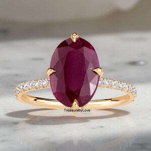 May include: A gold ring featuring a large, oval-cut ruby gemstone. The ruby is a deep red color and is the focal point of the ring. The band is adorned with small, sparkling diamonds. The ring is set against a neutral background.