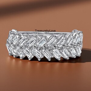 Puede incluir: Un anillo plateado con una serie de piedras preciosas claras talladas en baguette dispuestas en un patrón en zigzag. El anillo está sobre un fondo marrón. El texto "TreasurebyLove" es visible en el anillo.