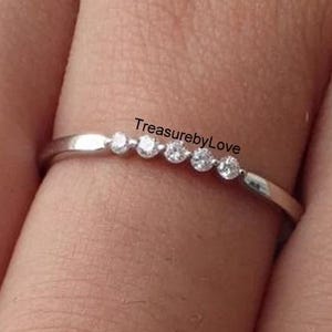 Pode incluir: Um anel de prata com cinco pequenas pedras preciosas redondas e claras dispostas em linha. O anel é fino e está em um dedo. O texto "TreasurebyLove" é visível.