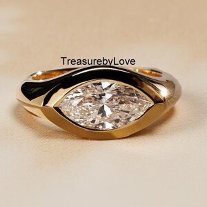 May include: A gold ring featuring a marquise-cut clear gemstone set in a smooth bezel setting. The band is polished gold with a thick, rounded profile. The text TreasurebyLove is printed in black above the ring on the surface.