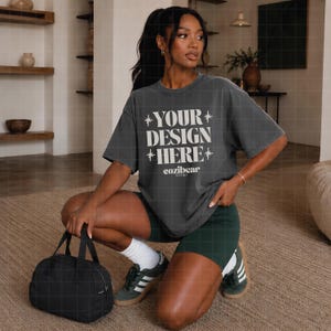 May include: A charcoal gray oversized t-shirt featuring the white text "YOUR DESIGN HERE" and "cozibear STUDIO" with star accents, paired with dark green athletic shorts, white crew socks, dark green sneakers, and a black duffel bag.