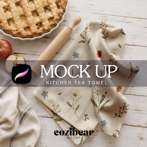 May include: A cream-colored kitchen tea towel featuring a repeating pattern of small blue flowers, white daisies, green stems, and red strawberries. The towel is displayed alongside a wooden rolling pin, a baked pie, and sliced apples.