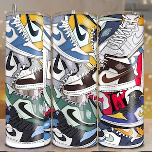 May include: Three stainless steel tumblers featuring a vibrant design of stacked trainers. The trainers are in various colours, including blue, yellow, brown, and green, with black accents. Each tumbler has a silver straw.