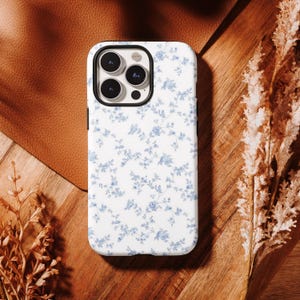 Può includere: Custodia per telefono bianca con un delicato motivo floreale blu. La custodia ha un bordo nero e un ritaglio per la fotocamera. La custodia del telefono è su una superficie di legno con una toppa in pelle e piante essiccate.