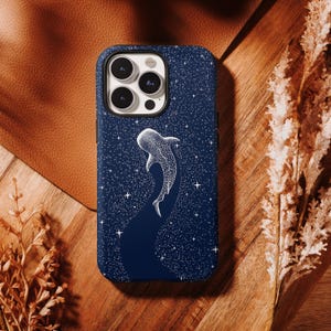 May include: A navy blue phone case featuring a white whale shark design against a starry night sky. The whale shark is depicted in a detailed, dotted style, with a dark blue trail. The case has a glossy finish and is set on a wooden surface.