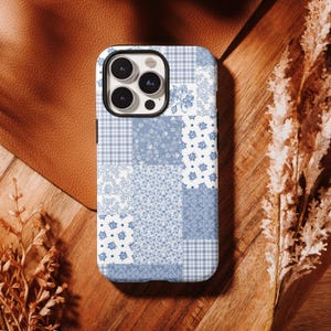 Puede incluir: Una funda de teléfono de retazos azul y blanco con una variedad de patrones, incluyendo cuadros, flores y diseños de cachemira. La funda tiene un marco negro para la lente de la cámara y está sobre una superficie de madera.