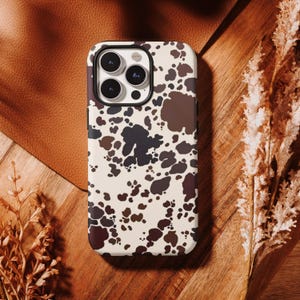 May include: A smartphone case with a cowhide pattern. The case features a cream-colored background with irregular brown and dark brown spots. The case has a black border and a camera cutout.