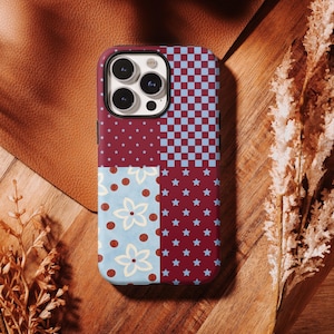 Puede incluir: Una funda de teléfono estampada con un diseño de patchwork. La funda está dividida en cuatro secciones con diferentes patrones: un campo burdeos con estrellas azules, un tablero de ajedrez azul y blanco, un campo azul claro con flores blancas y lunares marrones, y un campo burdeos con estrellas azules.