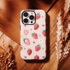 Könnte beinhalten: Eine Handyhülle mit rosa Hintergrund und einem Muster aus roten Erdbeeren, weißen Blumen und kleinen roten Herzen. Die Hülle hat einen schwarzen Rand um die Kameralinsen.