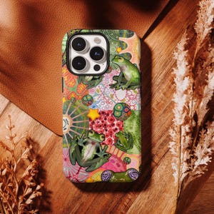 May include: A vibrant phone case with a patchwork design showcasing green frogs, stars, and diverse patterns. The case has a glossy finish and a camera cutout. The background is a wooden surface with a dried plant.