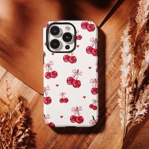 Op de afbeelding: Een witte telefoonhoes met een patroon van rode kersen met roze strikken en kleine rode hartjes. De hoes heeft een zwarte rand en een camera-uitsparing. Het ontwerp wordt herhaald.