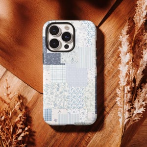Może przedstawiać: Etui na telefon z patchworkowym wzorem w odcieniach niebieskiego i białego. Etui posiada różne wzory, w tym nadruki kwiatowe, paski i kratę. Etui na telefon leży na drewnianej powierzchni z skórzanym tłem i suszonymi roślinami.