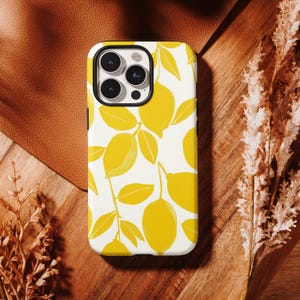 May include: A white iPhone case with a yellow lemon and leaf pattern. The case has a black border and a camera cutout. The design features yellow lemons and leaves on a white background. The case is on a wooden surface.