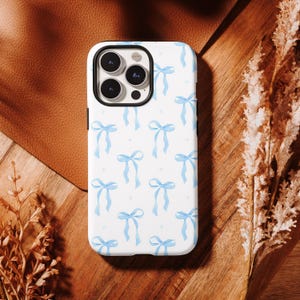 May include: White phone case featuring a pattern of light blue bows and small dots. The case has a black border. The bows are evenly spaced across the white surface, covering the back and sides of the phone.