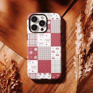 Puede incluir: Funda de teléfono con diseño de patchwork en rojo, blanco y gris. El diseño incluye lazos, cerezas, estrellas y motivos florales. Tiene un recorte para la cámara y está sobre una superficie de madera.