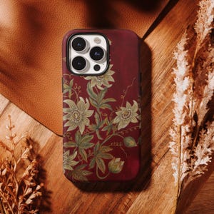 Pode incluir: Capa de telefone bordô com um design floral de folhas verdes e flores creme. A capa tem um acabamento brilhante e é feita de um material duro. A capa do telefone está em uma superfície de madeira.
