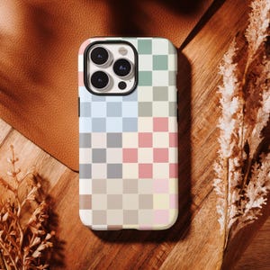 Op de afbeelding: Een telefoonhoesje met een kleurrijk ruitpatroon in pastelkleuren blauw, roze, groen en beige. De hoes heeft een zwart cameramodule en ligt op een houten oppervlak.