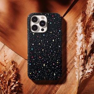May include: Black phone case with a colorful starry night design. The case features a pattern of white dots, stars, and crescent moons in various colors. The phone case is on a wooden surface.