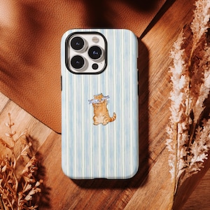 May include: A phone case with a watercolour illustration of a ginger cat wearing spectacles and holding a fish. The case has a light blue and white striped background. The phone case is on a wooden surface.