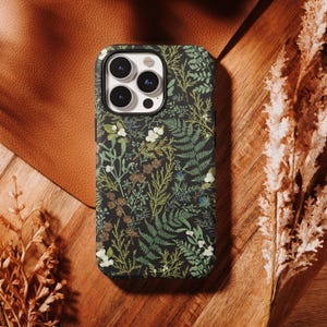 May include: A black phone case with a botanical pattern featuring green ferns, white flowers, and brown accents. The case is on a wooden surface with dried plants and a leather backdrop.
