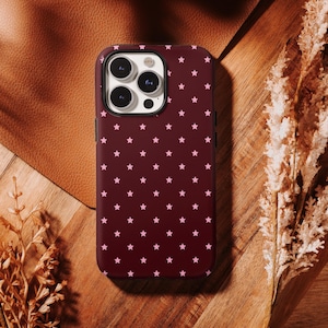 May include: A burgundy phone case with a pattern of small pink stars. The case has a black border and a camera cutout. The phone case is on a wooden surface with a leather background and dried plants.