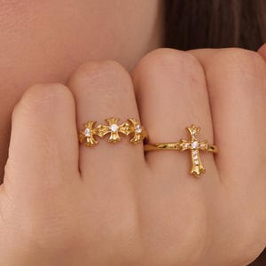 Könnte beinhalten: Zwei goldene Ringe mit Kreuz- und Blumenmotiven. Ein Ring hat ein Kreuzdesign mit kleinen, klaren Steinen. Der andere Ring hat ein Blumenmuster mit kleinen, klaren Steinen.