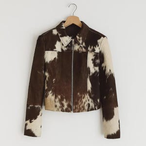 May include: A brown and white cowhide jacket with a zip-up front and a collared neckline. The jacket is hanging on a wooden hanger. The jacket's design features a mix of brown and white patches.