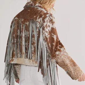 Puede incluir: Chaqueta de piel de vaca marrón y blanca con flecos grises. La chaqueta tiene cuello, mangas largas y largo recortado. Los flecos añaden un toque elegante. Es una prenda llamativa.