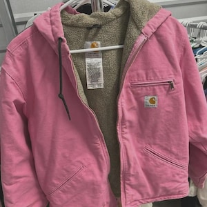 May include: A pink Carhartt hooded jacket with a zip-up front and multiple pockets. The jacket has a tan-colored, fuzzy lining and a black drawstring on the hood. The Carhartt logo is visible on the chest.