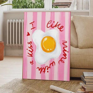 May include: A framed print with a fried egg illustration on a pink striped background. Red cursive text reads "I like mine with a kiss" around the egg. The artwork is displayed on a wooden floor.