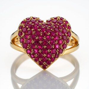 May include: A gold ring featuring a heart-shaped design, densely covered with small, round, deep red gemstones. The ring has a split band and a polished, reflective surface.