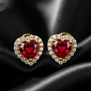 May include: A pair of heart-shaped stud earrings. Each earring has a vibrant red gemstone centre, encircled by a halo of small, sparkling white stones. The setting is gold-toned, and the earrings are set against a black background.