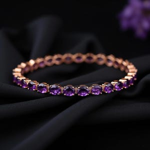 May include: A rose gold bracelet with oval-cut purple gemstones. The bracelet is set on a black background, highlighting the sparkle of the gemstones. A beautiful piece of jewelry.