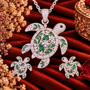 May include: A jewellery set featuring a turtle pendant on a chain and matching turtle earrings. Each piece is crafted in a metallic tone, encrusted with clear white stones and accented with marquise-cut green stones in a floral pattern.