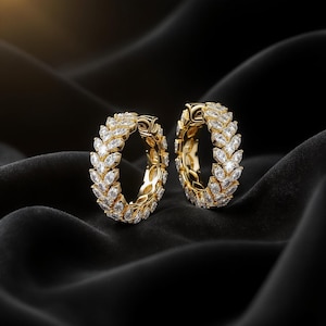 May include: Two gold hoop earrings, each encrusted with numerous clear, marquise-cut gemstones. The earrings are set against a black, silky background, creating a luxurious and elegant aesthetic. The gemstones are arranged in a leaf-like pattern.