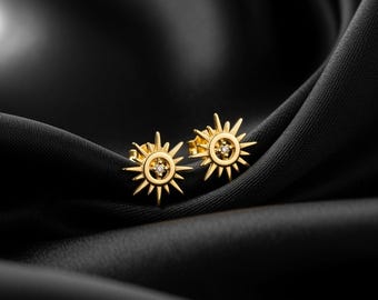 Peut inclure: Une paire de boucles d'oreilles à clous dorées en forme de soleil, chacune ornée d'une petite pierre précieuse transparente au centre. Les boucles d'oreilles sont présentées sur un tissu noir et soyeux.