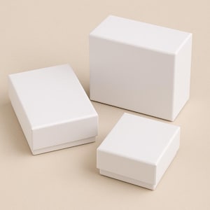 Op de afbeelding: Drie witte rechthoekige geschenkdozen van verschillende formaten zijn gerangschikt op een lichtbeige oppervlak. De dozen zijn allemaal gesloten, met strakke lijnen en een matte afwerking. De dozen zijn waarschijnlijk voor sieraden of kleine cadeaus.