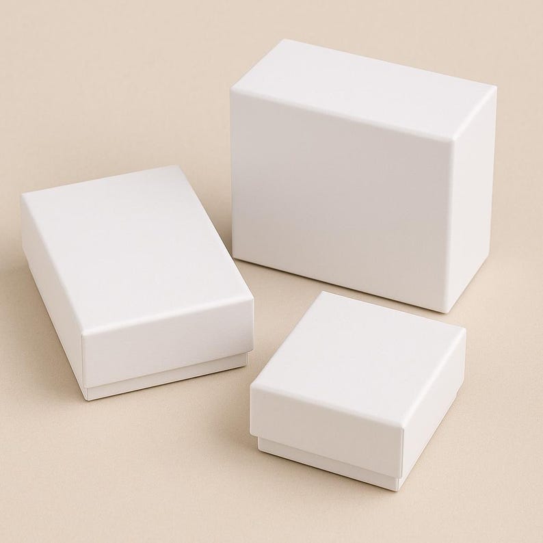Op de afbeelding: Drie witte rechthoekige geschenkdozen van verschillende formaten zijn gerangschikt op een lichtbeige oppervlak. De dozen zijn allemaal gesloten, met strakke lijnen en een matte afwerking. De dozen zijn waarschijnlijk voor sieraden of kleine cadeaus.