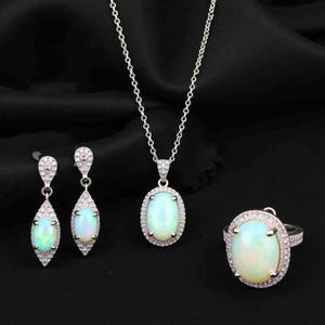 May include: A jewellery set comprising a silver necklace, earrings, and ring. Each item features an oval, opalescent stone encircled by small, clear stones. The earrings are drop style, and the ring has a wide band.