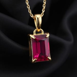 May include: A gold pendant necklace featuring a rectangular, emerald-cut deep red gemstone held by four prongs. The pendant is suspended from a matching gold chain, showcasing a polished metal finish and a vibrant, translucent red stone.