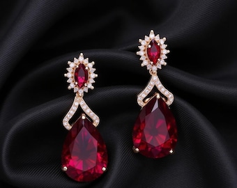 May include: A pair of gold-toned drop earrings with large, teardrop-shaped red gemstones. The earrings feature smaller red stones surrounded by clear crystals at the top and along the connecting accents, set against a black background.