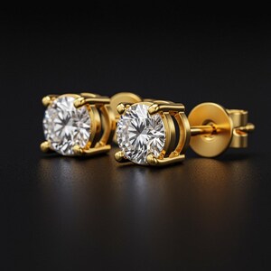 May include: A pair of gold stud earrings with a round, clear gemstone. The earrings have a classic design with a four-prong setting and a post back. The gold has a polished finish, and the gemstones sparkle against the dark background.