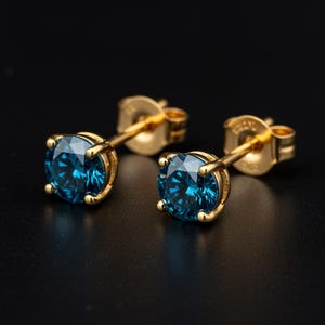 May include: A pair of gold stud earrings with round, faceted, teal-colored gemstones. The earrings are set in a four-prong setting and have a post and butterfly clasp. The background is black.