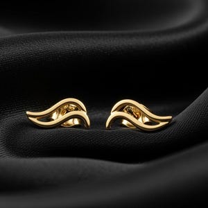 May include: A pair of gold-colored earrings with a flowing, abstract design. The earrings are displayed on a black satin fabric, creating a contrast that highlights their shape and shine. The earrings have a delicate, openwork structure.